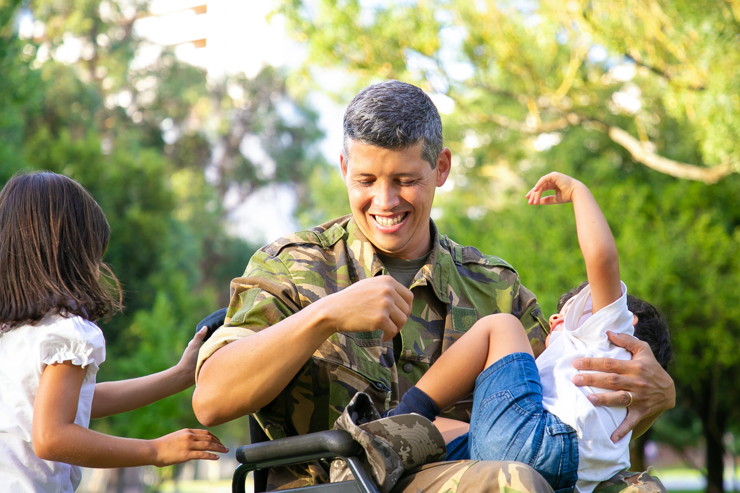 Cheerful disabled military dad enjoying leisure time with two kids in park Girl holding wheelchair handles boy resting on dads lap Veteran of war or disability concept